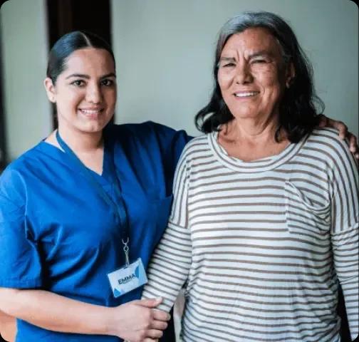 The image shows a healthcare professional in a blue uniform with a name tag, smiling and supporting an elderly person who is also smiling. The elderly person is dressed in a striped shirt, and they appear to be indoors in a well-lit setting, demonstrating a friendly and supportive relationship